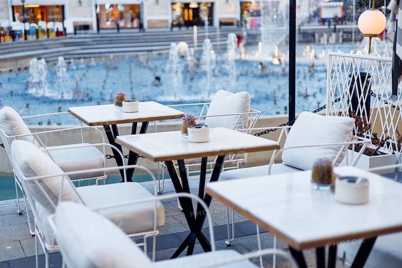 Cozy tables and chairs set near a beautiful fountain in a lively urban environment. The soft glow of lights reflects off the water as people enjoy the evening ambiance