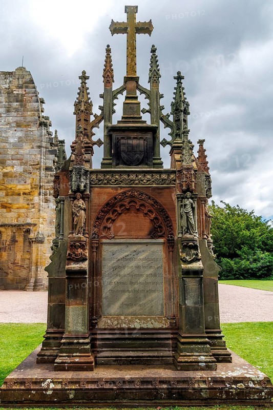 A Gothic Revival memorial cross and carved monument stand in the grounds of 15th century Rosslyn Chapel, Scotland, United Kingdom