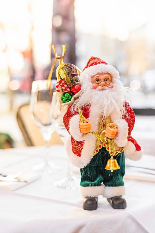 A decorative Santa Claus figurine stands on a table. The Santa has a white beard, red suit, and holds bells and a sack. The background is softly blurred