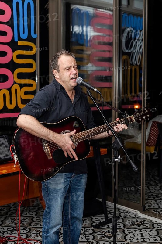 A middle-aged Caucasian man with short brown hair sings and plays an acoustic guitar in a vibrant cafe setting. Colorful decor and a lively atmosphere surround him