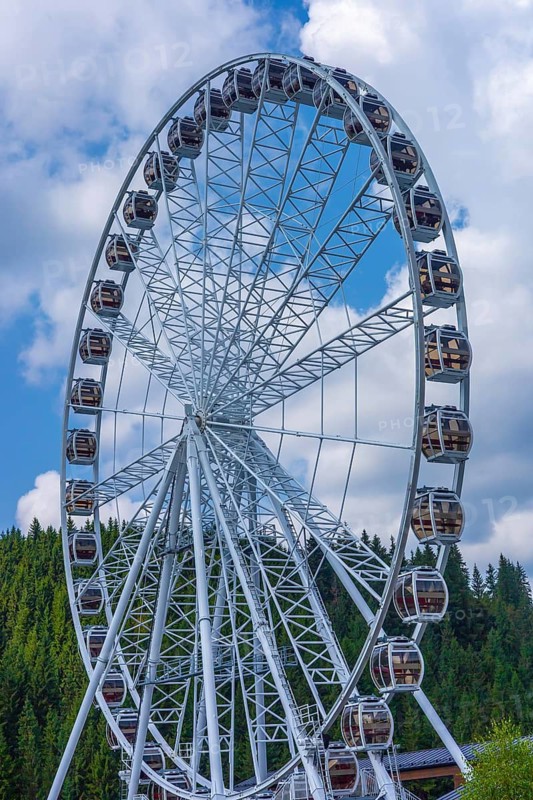 A large Ferris wheel stands against a blue sky with fluffy clouds. The structure is metallic and surrounded by green trees