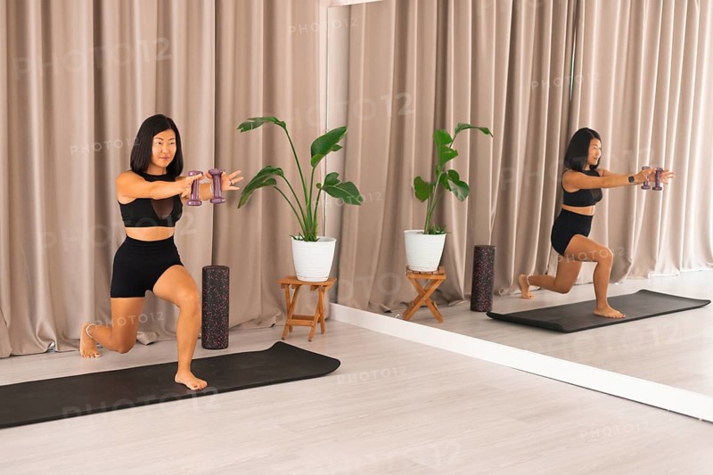 A woman performs lunging exercises with dumbbells indoors, surrounded by plants and mirrors