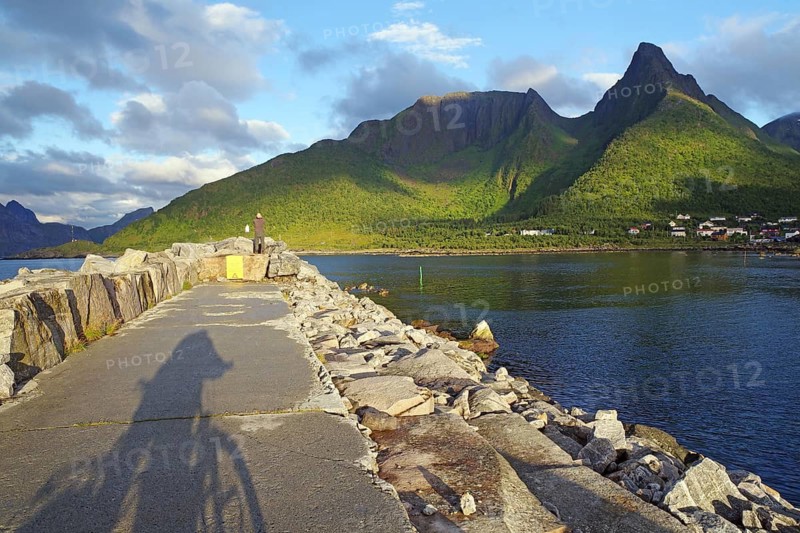 A stone path along the sea with a bicycle shade. The view leads to green mountains, partly sunny