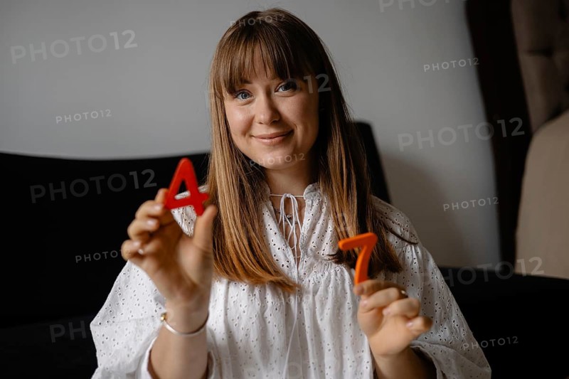 A woman indoors holds red numbers 4 and 7, smiling casually