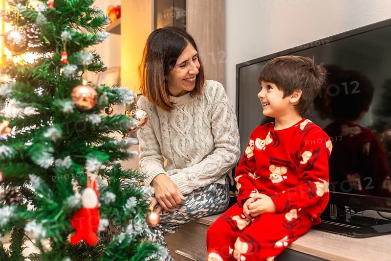 Mother and young son in pajamas sharing joyful smiles and warm moments by a decorated christmas tree at home, enjoying cozy, candid holiday bonding and laughter