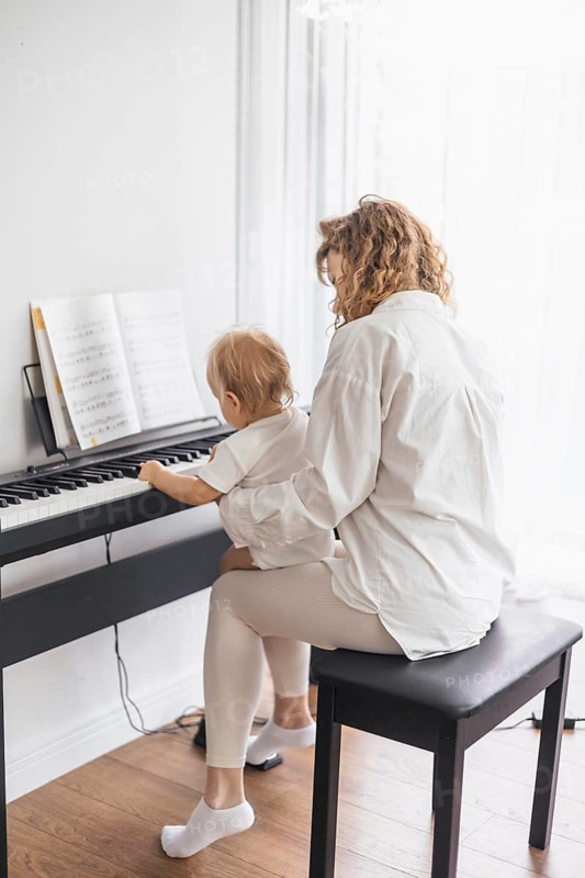 A woman and child sit at a piano from the backside, in a peaceful and bright room