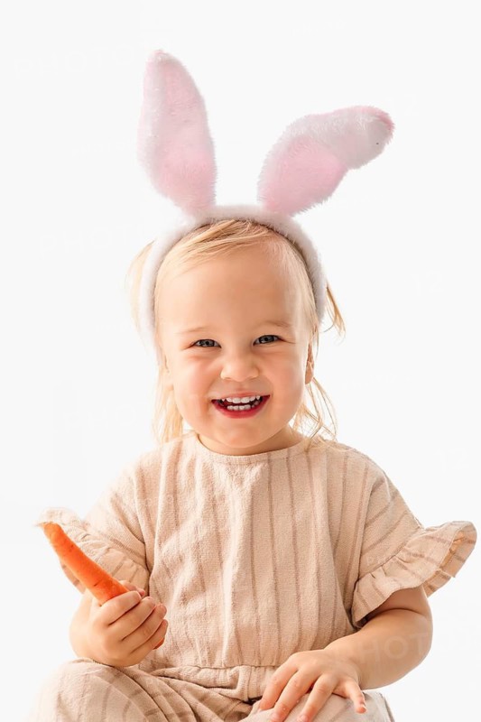 A young girl wearing bunny ears smiles joyfully while holding a carrot. She participates in Easter celebrations. This fun moment captures the spirit of the holiday and family traditions