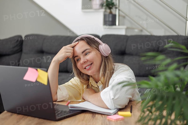 Young blond European woman with headphones lying on floor near laptop and notebook in modern living room