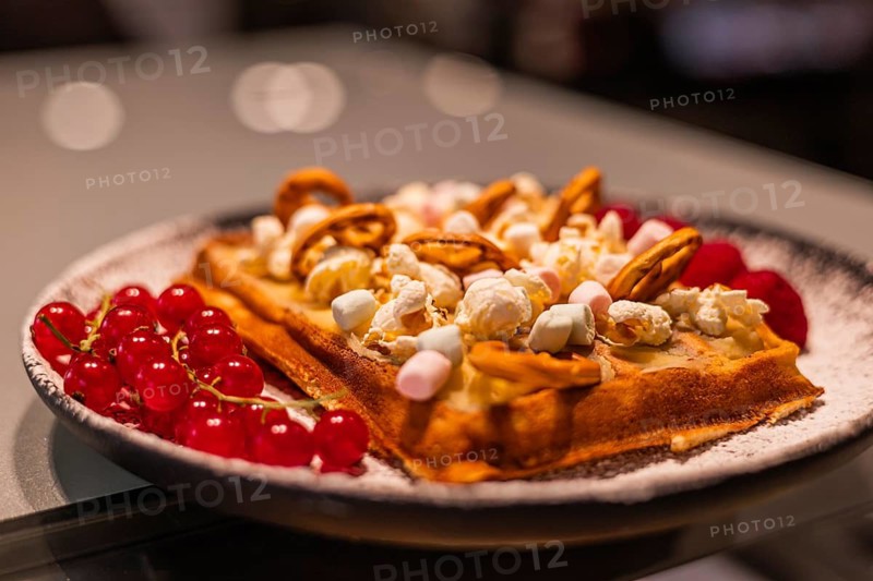 A delicious waffle topped with popcorn, marshmallows, and nuts, served with fresh red berries on a black plate