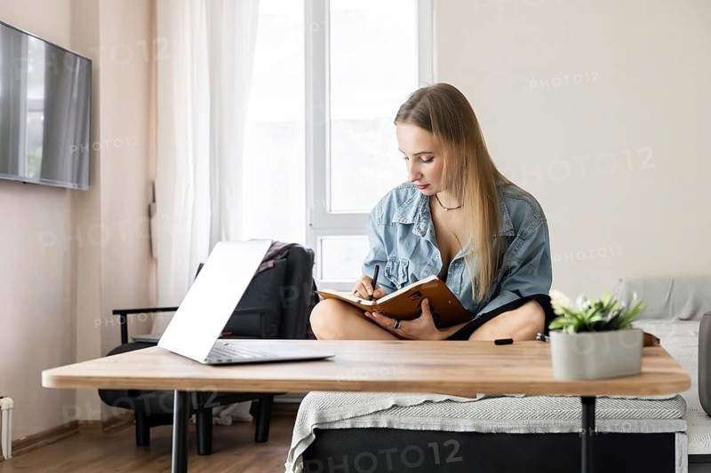 Young Caucasian woman with long blond hair sits cross-legged at a table, writing in a notebook. A laptop and a potted plant are on the table. Bright indoor setting