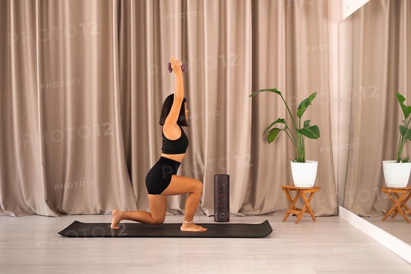 A woman lunges with dumbbells inside a minimalistic room with plants and a curtain