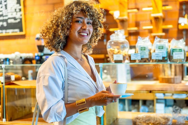 Smiling woman with curly hair holding a cup of coffee, standing in a vibrant new york city cafe, representing urban lifestyle and casual moments
