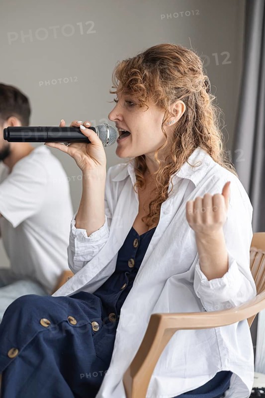 A young Caucasian woman with curly hair sings into a microphone at a home concert. A man is seated nearby, creating a cozy family atmosphere