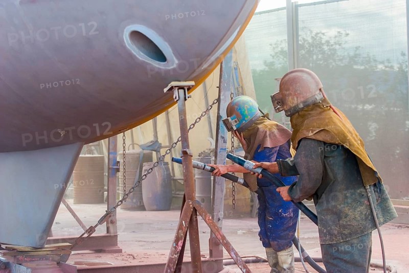 Worker sandblasting a sailing yacht hull in full protective clothing and helmet, creating a cloud of abrasive dust. Whangarei, Northland, New Zealand