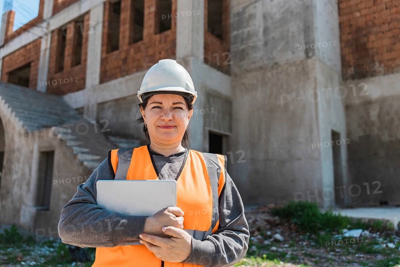 Woman stands on a construction site. She wears a hard hat and an orange safety vest, holding a tablet. The building is in progress behind her