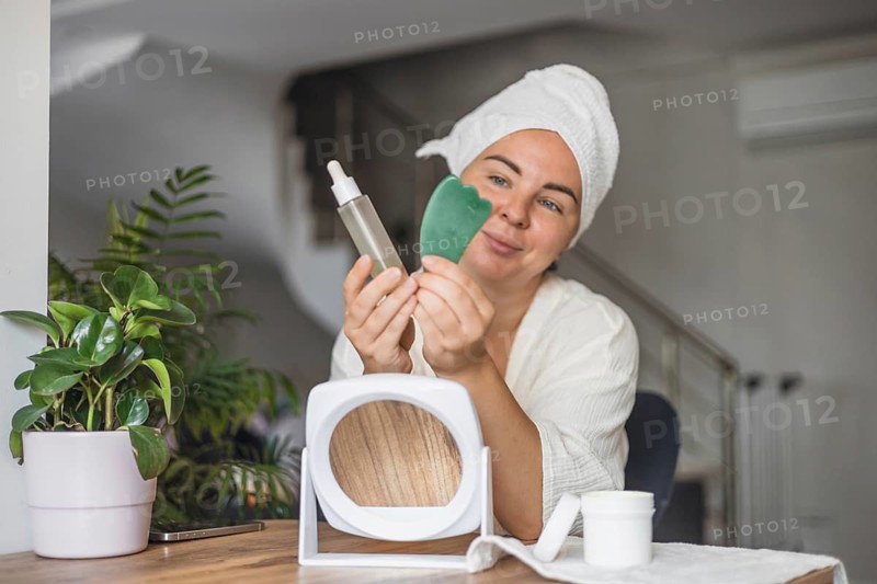 A young Hispanic woman with a towel on her head holds a gua sha tool and a serum bottle. A mirror and a plant are on the table, creating a serene beauty setting