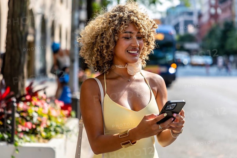 Young woman with curly hair smiling and looking at her smartphone, standing on a bustling street in new york city, enjoying urban lifestyle and staying connected