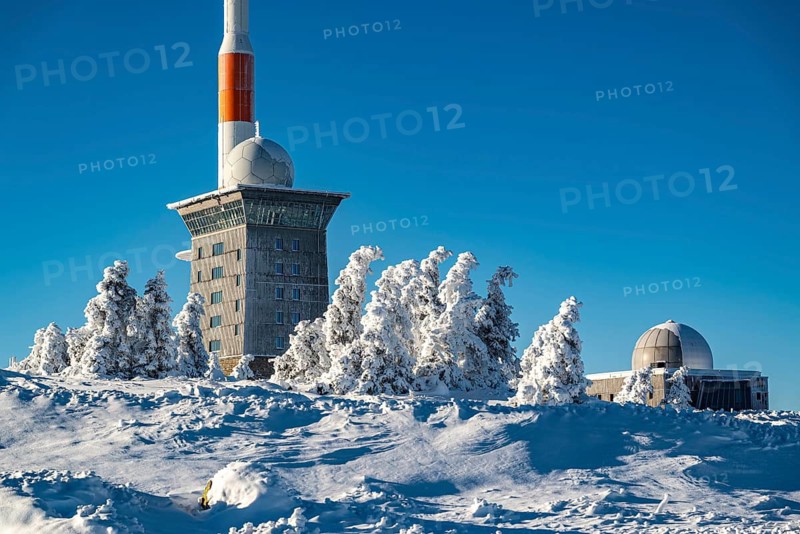 View over snow-covered spruce trees of the icy Brocken Hotel and the telecommunications tower on the summit of Mount Brocken, on a sunny winter day under clear blue skies, Harz National Park, Saxony-Anhalt, Germany