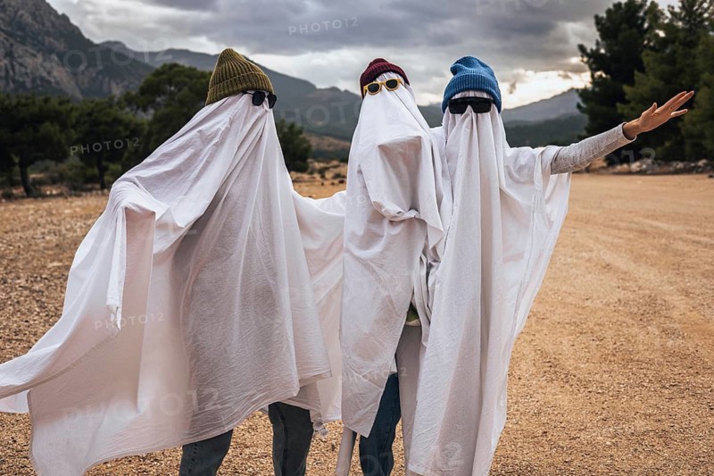 Three people dressed as ghosts for Halloween. They wear white sheets and sunglasses, standing outdoors with mountains in the background