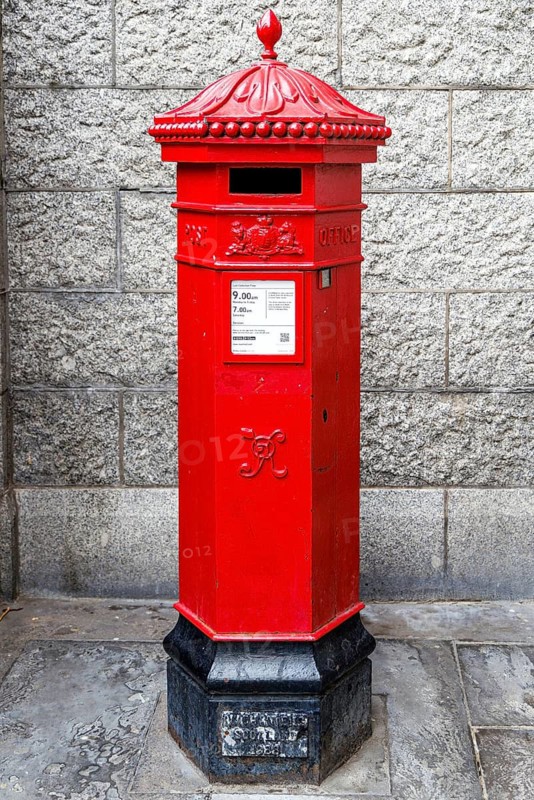 A hexagonal red pillar post box with an ornate crown finial and royal cypher stands against a granite block wall, a design tracing back to the Victorian introduction of Britain's first pillar boxes in 1852 and still a familiar sight on London streets, London, England, United Kingdom