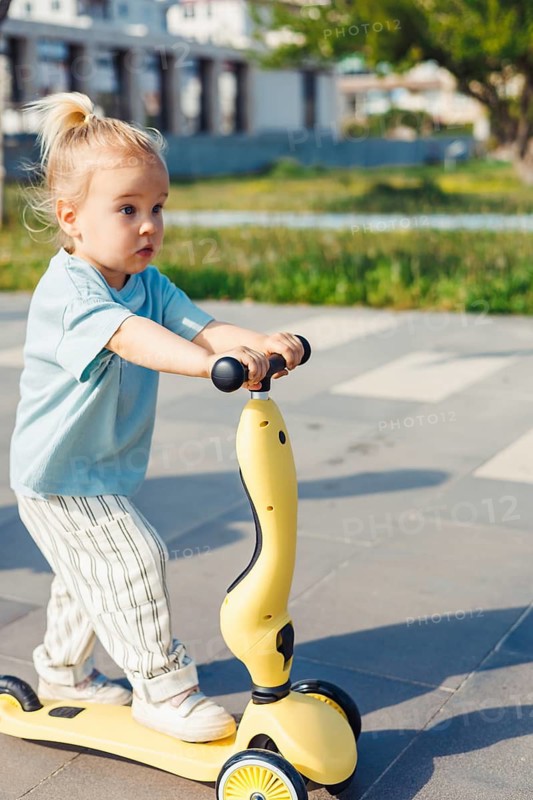 A young child with a ponytail rides a yellow scooter on a paved path in a park. Surrounding greenery and playground equipment create a cheerful atmosphere on this bright day