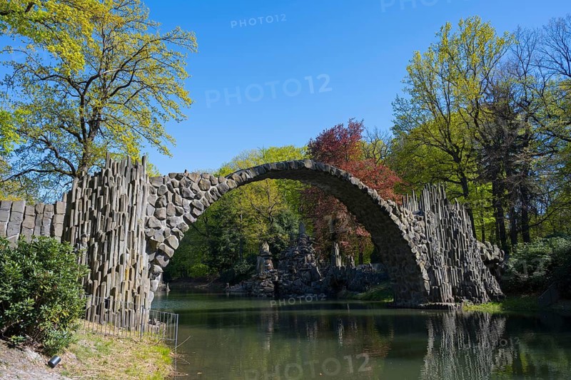 Stone bridge against colorful, wooded background on a sunny day, over a calm body of water, Rakotz Bridge, Rakotzsee, arched bridge, reflection, basalt organ, Kromlau, Görlitz district, Saxony, Germany