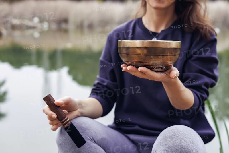 A young Caucasian woman with long brown hair holds a wooden bowl near a serene lake. Trees and greenery are visible in the background