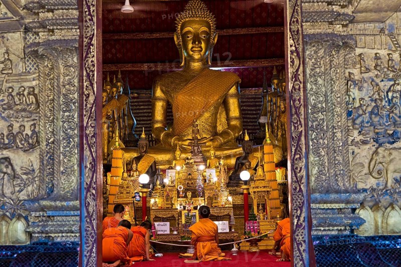 Buddhist monks and novices sit in front of a large Buddha statue in Wat May Souvannapoumaram, Luang Prabang, Laos, Southeast Asia
