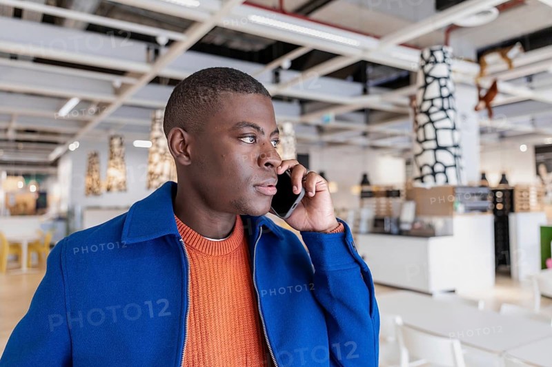 A young African man with short hair is talking on a smartphone in a modern interior space. The setting features contemporary furniture and decor