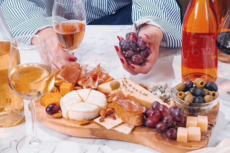 A group of friends gathers around a table enjoying drinks and a platter filled with cheese, meats, olives, and grapes. The atmosphere is lively and casual during the afternoon