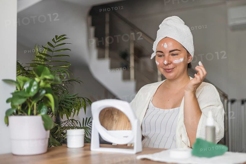 A woman in a towel applies cream to her face while seated at a wooden table
