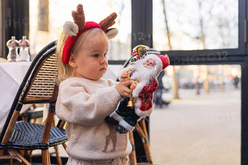 A young Caucasian girl with blonde hair wears a reindeer headband. She holds a Santa Claus toy in a festive setting, celebrating Christmas and New Year