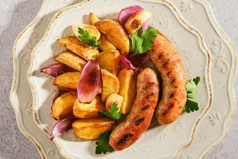 Two grilled sausages sit beside roasted potatoes and pieces of onion on decorative plate. Fresh parsley garnishes the dish. This meal appears ready to eat and is visually appealing