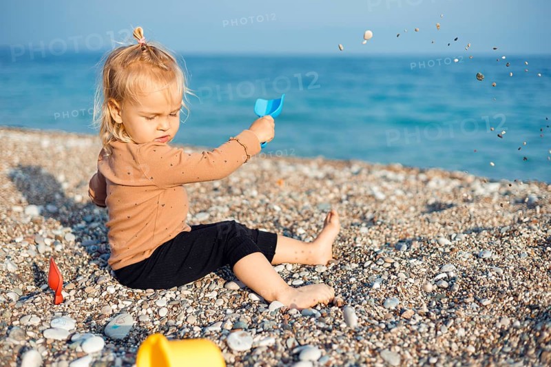 A joyful child sits on a sunny beach, happily playing with sand and pebbles. The clear blue sea sparkles in the background, while the child explores the sandy texture with delight