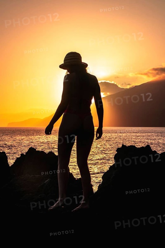 Woman wearing a straw hat and bikini standing on volcanic rocks, silhouetted against a golden sunset over the ocean on el hierro island in the canary islands, enjoying a peaceful summer vacation