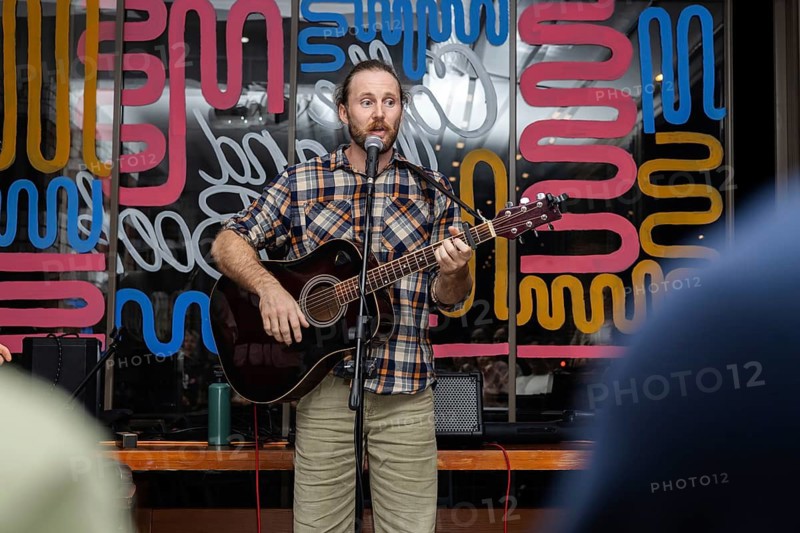A young Caucasian man with a beard plays an acoustic guitar while singing on stage. Colorful abstract background enhances the lively atmosphere