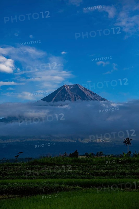 A majestic volcano rises above a green landscape surrounded by clouds under a clear blue sky, Volcano, Sumatra, Jakarta, Indonesia