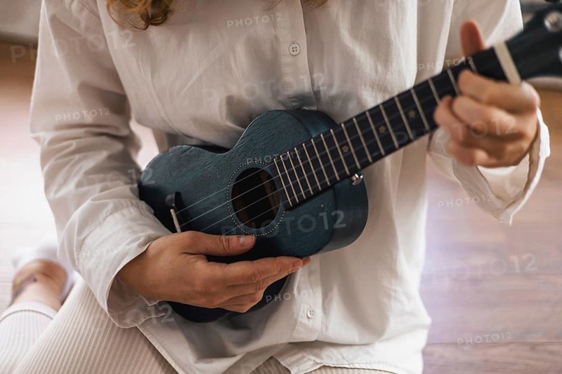 Person in white shirt playing a ukulele while seated on a wooden floor