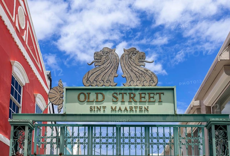 Colorful decorative Old Street Sint Maarten entrance gate with twin lion sculptures and ornate green ironwork in Philipsburg, flanked by colorful colonial buildings under a blue sky