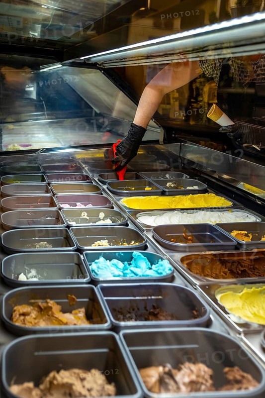 Hand wearing black glove preparing to scoop colorful gelato from a display case, showing a variety of sweet frozen dessert flavors in metal trays at a shop counter