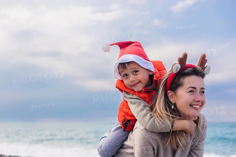 A mother and her young son enjoy a winter day at the beach. The mother has brown hair and wears a cozy sweater. The boy wears a red Santa hat and an orange jacket