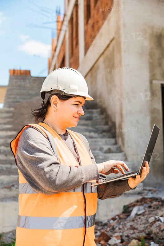 Woman in a hard hat and orange vest uses a laptop on a construction site. Stairs and an unfinished doorway are visible in the background
