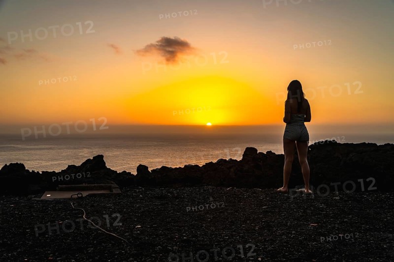 Woman standing on the volcanic rocky terrain watching a vibrant orange sunset over the atlantic ocean, finding peace and tranquility during a summer vacation on the island of el hierro