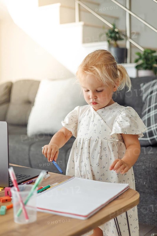 Young blond European girl drawing with colored pencils at home near laptop and sofa in living room