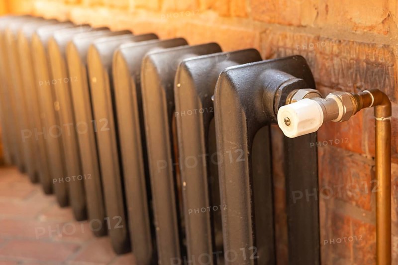 A traditional retro metallic radiator is connected to a brass pipe in a warm and inviting interior setting. The radiator features a classic design and is positioned against a rustic exposed brick wall, adding character to the room. This heating element is essential for providing warmth during winter months