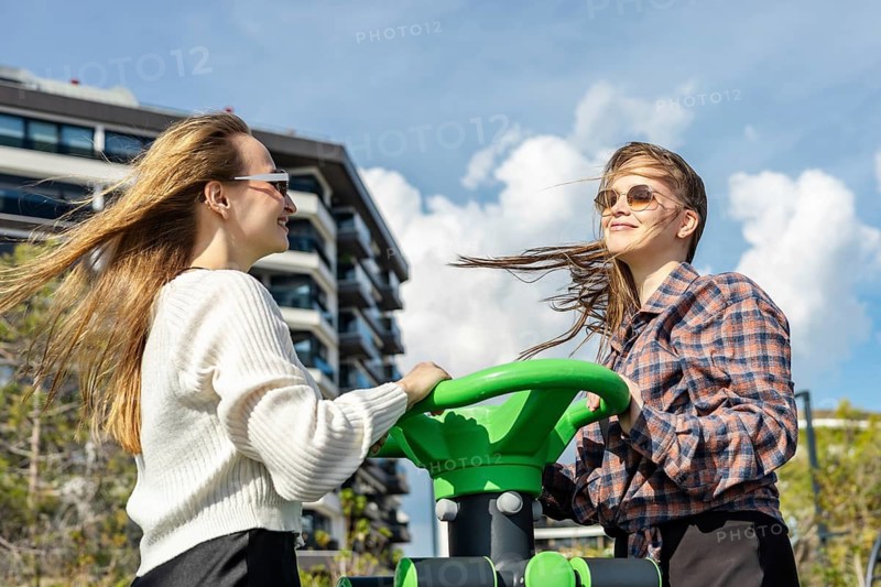 Two young women engage in outdoor exercise at a fitness station in an urban park. One has long brown hair, the other has short blond hair. Both wear casual clothing