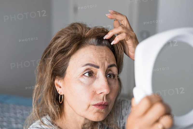 Mature woman inspects her appearance in mirror, looking at her gray roots hair. Natural light fills room, creating calm and personal atmosphere. age-related changes, naturalness and natural aging