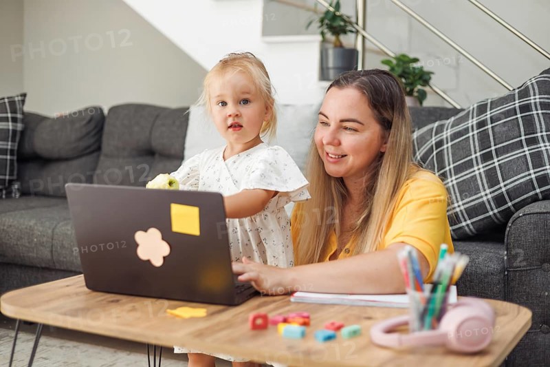 Young Caucasian woman and blond Caucasian girl using laptop at home table with school supplies and apple, online learning concept