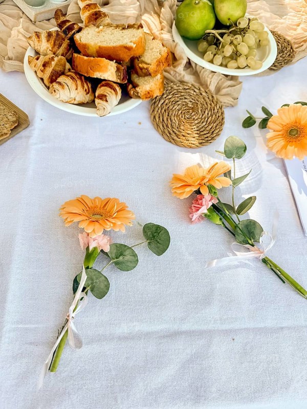 A table setting with fresh flowers, bread, and fruits. The scene includes orange flowers, green apples, and grapes on a white tablecloth
