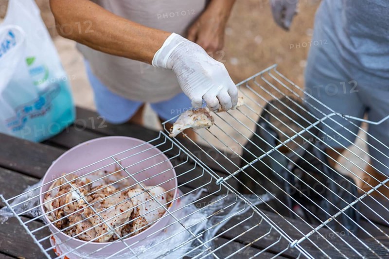 A person with brown skin and gloves prepares food on a grill. A bowl of marinated meat is nearby. The setting is outdoors, suggesting a barbecue or picnic atmosphere